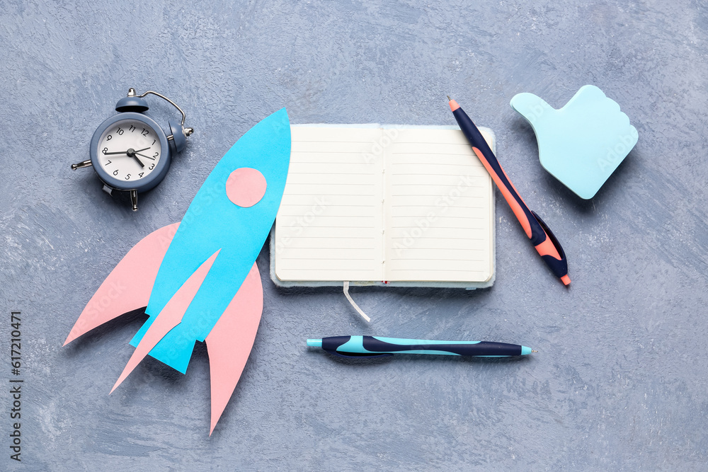 Blue paper rocket with open notebook, pens and alarm clock on grey grunge background