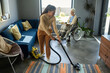 © AnnaStills - Youthful girl with vacuum cleaner helping her grandmother with disability with domestic chores while senior woman reading book by window
