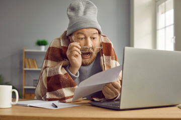 Stressed elderly man in warm clothes and glasses sits at desk at home in cold apartment, holds domestic utility bill, reads information, looks at figures shocked by expensive Internet or heating costs