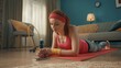 © kinomaster - A young woman in a pink tracksuit and sports headband is lying on a sports mat on the floor. A young woman leaning on her elbows looks into the phone. Close up. Sports video lessons.