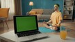 © kinomaster - Laptop with green screen and a glass of juice on the table in the living room. In the background, a man in a tracksuit and sports headband sits in a lotus position. Sports concept. Yoga class