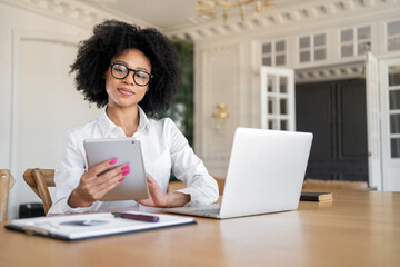 A woman works on the Internet on a computer in an office smart with glasses freelancer lawyer manager assistant uses a tablet stands and looks at the camera smiling.