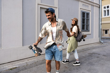 Tourists couple skateboarders riding skates. Happy young travelers longboarding together on sunset on pavement. Stylish man and woman in trendy outfit skating outdoors in touristic destination streets