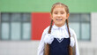 © lenblr - Junior schoolgirl with long braids stands with excited and smiling expression against modern school. Brown-haired girl enjoys going to school