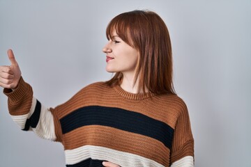 Poster - Young beautiful woman wearing striped sweater over isolated background looking proud, smiling doing thumbs up gesture to the side