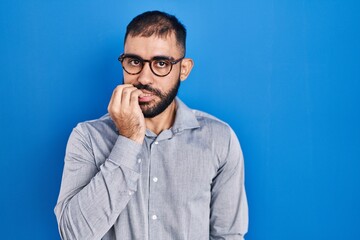 Sticker - Middle east man with beard standing over blue background looking stressed and nervous with hands on mouth biting nails. anxiety problem.