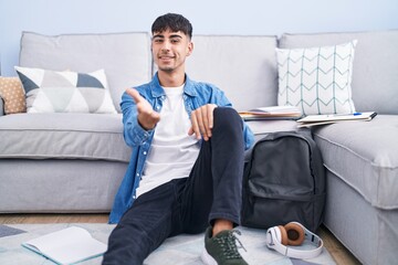 Sticker - Young hispanic man sitting on the floor studying for university smiling cheerful offering palm hand giving assistance and acceptance.