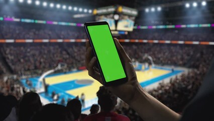 Basketball Championship: Person's Hand Holding a Smartphone with a Green Screen Display. Sports Fans in an Arena Cheering for a Team to Win. Template to Use for Social Media, Scores, Results, Betting