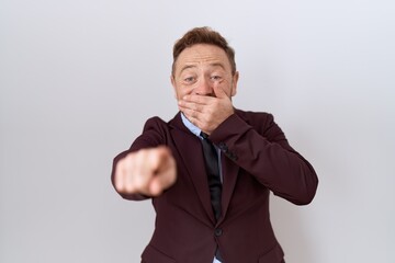 Middle age business man with beard wearing suit and tie laughing at you, pointing finger to the camera with hand over mouth, shame expression