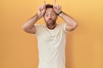 Poster - Middle age man with beard standing over yellow background doing funny gesture with finger over head as bull horns