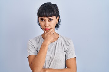 Sticker - Young hispanic woman wearing casual t shirt over blue background looking confident at the camera with smile with crossed arms and hand raised on chin. thinking positive.