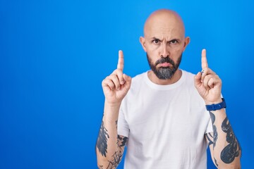 Wall Mural - Hispanic man with tattoos standing over blue background pointing up looking sad and upset, indicating direction with fingers, unhappy and depressed.