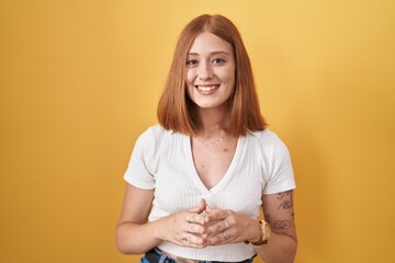 Wall Mural - Young redhead woman standing over yellow background hands together and fingers crossed smiling relaxed and cheerful. success and optimistic