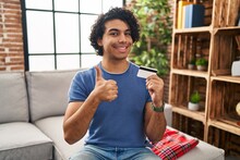 Hispanic Man With Curly Hair Holding Credit Card Smiling Happy And Positive, Thumb Up Doing Excellent And Approval Sign