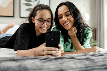 Wall Mural - Mother and teenage daughter using smartphone together lying in bed at home. Latina mother and daughter looking at phone watching social media videos while relaxing in bedroom. family and technology