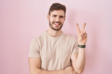 Poster - Hispanic man with beard standing over pink background smiling with happy face winking at the camera doing victory sign with fingers. number two.