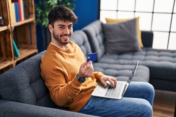 Wall Mural - Young hispanic man using laptop and credit card sitting on sofa at home