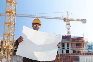 civil engineer architect foreman construction worker on construction site with crane and building shell in background holding plan drawing 