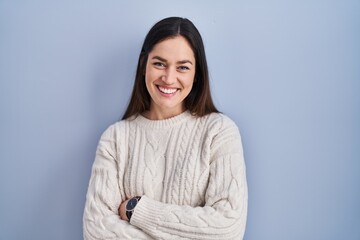 Poster - Young brunette woman standing over blue background happy face smiling with crossed arms looking at the camera. positive person.