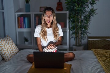 Wall Mural - Young blonde woman sitting on the bed at home watching a movie from laptop in shock face, looking skeptical and sarcastic, surprised with open mouth