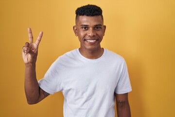 Wall Mural - Young hispanic man standing over yellow background showing and pointing up with fingers number two while smiling confident and happy.