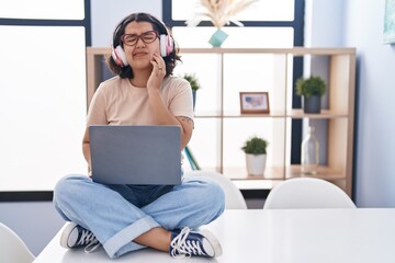 Poster - Young hispanic woman using laptop sitting on the table wearing headphones touching mouth with hand with painful expression because of toothache or dental illness on teeth. dentist