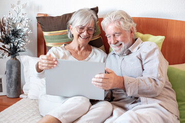 Wall Mural - Cheerful senior white-haired couple resting in bed using wireless technology with laptop. Elderly man and woman enjoying relax looking at social media