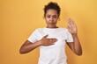 Young hispanic woman with curly hair standing over yellow background swearing with hand on chest and open palm, making a loyalty promise oath