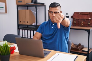 Wall Mural - Young hispanic man working at the office with laptop looking unhappy and angry showing rejection and negative with thumbs down gesture. bad expression.