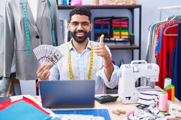 Poster - Hispanic man with beard dressmaker designer holding dollars smiling happy and positive, thumb up doing excellent and approval sign