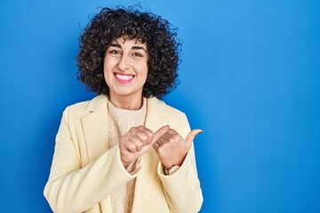 Wall Mural - Young brunette woman with curly hair standing over blue background pointing to the back behind with hand and thumbs up, smiling confident