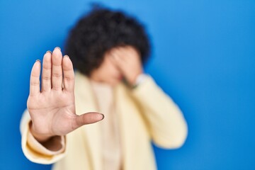 Sticker - Young brunette woman with curly hair standing over blue background covering eyes with hands and doing stop gesture with sad and fear expression. embarrassed and negative concept.