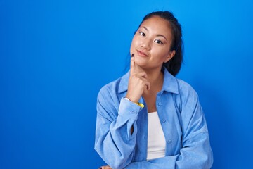 Poster - Asian young woman standing over blue background with hand on chin thinking about question, pensive expression. smiling with thoughtful face. doubt concept.