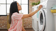 Young beautiful hispanic woman turning on washing machine at laundry room