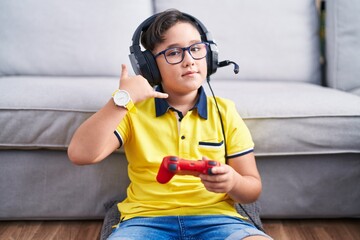 Poster - Young hispanic kid playing video game holding controller wearing headphones smiling doing phone gesture with hand and fingers like talking on the telephone. communicating concepts.