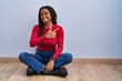 © Krakenimages.com - Young african american with braids sitting on the floor at home cheerful with a smile on face pointing with hand and finger up to the side with happy and natural expression