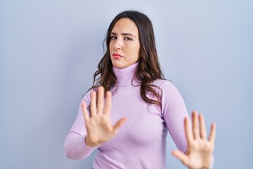 Poster - Young brunette woman standing over blue background moving away hands palms showing refusal and denial with afraid and disgusting expression. stop and forbidden.