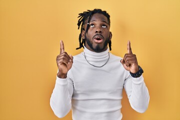 Wall Mural - African man with dreadlocks wearing turtleneck sweater over yellow background amazed and surprised looking up and pointing with fingers and raised arms.