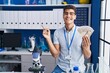 © Krakenimages.com - Young hispanic man working at scientist laboratory holding dollars smiling happy pointing with hand and finger to the side