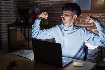 Poster - Young hispanic man with beard working at the office at night showing arms muscles smiling proud. fitness concept.