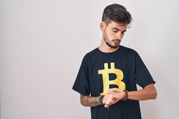 Poster - Young hispanic man with tattoos wearing bitcoin t shirt checking the time on wrist watch, relaxed and confident