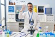 © Krakenimages.com - Young hispanic man with beard working at scientist laboratory smiling positive doing ok sign with hand and fingers. successful expression.