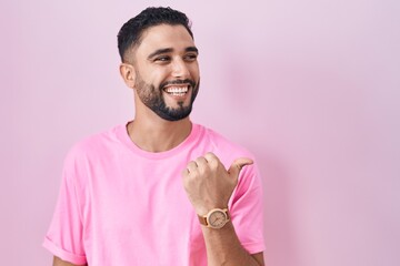 Hispanic young man standing over pink background smiling with happy face looking and pointing to the side with thumb up.