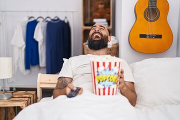 Canvas Print - Young hispanic man with beard and tattoos eating popcorn in the bed smiling and laughing hard out loud because funny crazy joke.