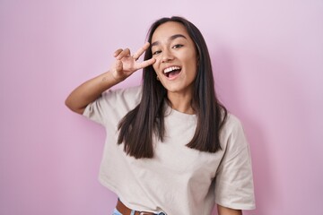Wall Mural - Young hispanic woman standing over pink background doing peace symbol with fingers over face, smiling cheerful showing victory