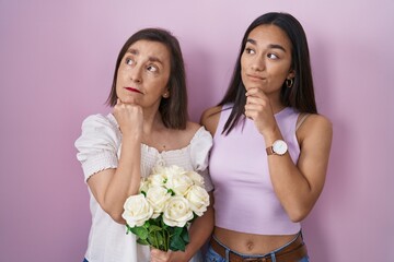 Wall Mural - Hispanic mother and daughter holding bouquet of white flowers with hand on chin thinking about question, pensive expression. smiling with thoughtful face. doubt concept.