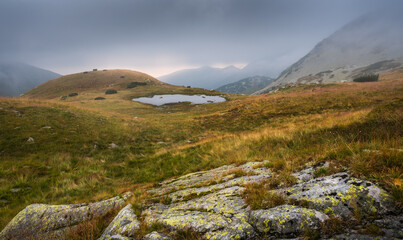 Wall Mural - Foggy Mountain Landscape with a Tarn and a Rock in Foreground at Sunset