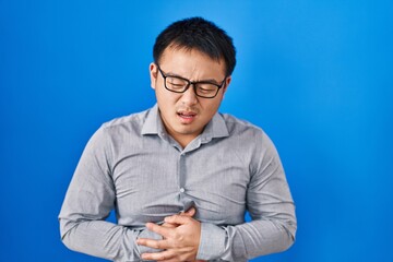 Canvas Print - Young chinese man standing over blue background with hand on stomach because indigestion, painful illness feeling unwell. ache concept.