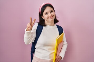 Canvas Print - Woman with down syndrome wearing student backpack and holding books smiling looking to the camera showing fingers doing victory sign. number two.