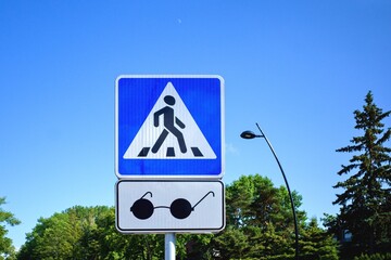 Road sign - Pedestrian crossing. A square blue and white metal plate with white stripes and a walking person. Transition for the blind with sound notification. Blue sky background.